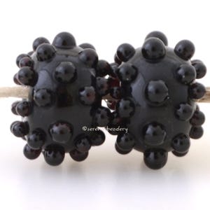 May include: Two dark black glass beads with a textured surface of small, round bumps. The beads are displayed on a neutral-colored string against a white background. The beads are likely for jewelry making.