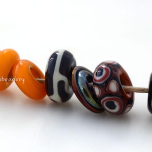 May include: A string of assorted glass beads on a light-colored cord. The beads include solid black, bright orange, black and white patterned, iridescent, multi-colored circular designs, and a white bead with green and blue accents. Text reads serenasbeadery.