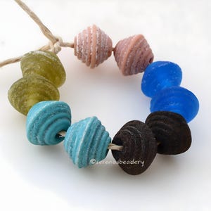 May include: A strand of colorful, textured beads in shades of green, pink, blue, and brown. The beads are strung on a natural-colored cord, creating a circular shape. The beads have a unique, ridged texture.