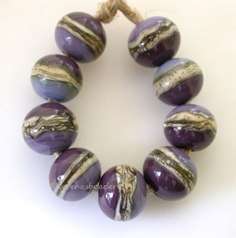Puede incluir: Una hebra de cuentas de vidrio de color morado y blanco con un patr&oacute;n de remolino. Las cuentas est&aacute;n enhebradas en un cord&oacute;n y est&aacute;n dispuestas en c&iacute;rculo.