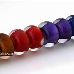 May include: A string of colorful glass beads in a gradient of colors, from brown to red, purple, and blue. Each bead is round and encased in clear glass, strung on a thin, natural-colored cord. The beads are likely for jewelry making.