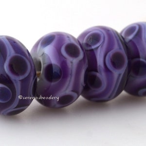 May include: Four purple glass beads with white swirls and dark purple spots. The beads are strung on a white cord.