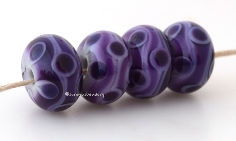 May include: Four purple glass beads with white swirls and dark purple spots. The beads are strung on a white cord.