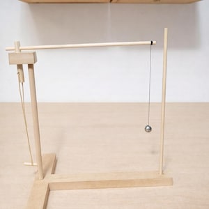 May include: A wooden Newton's cradle toy with a light-colored finish. The cradle features a horizontal beam, vertical supports, and a hanging metal ball. The design is simple and geometric, with a focus on the interplay of lines and shapes.