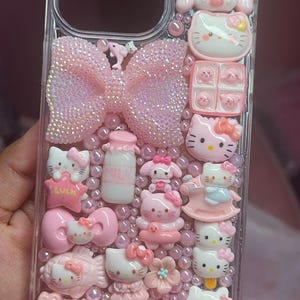Può includere: Una custodia per telefono trasparente decorata con personaggi Hello Kitty rosa, fiocchi e altri ciondoli. La custodia è piena di glitter e perline rosa.