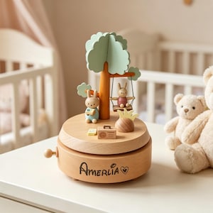 Peut inclure: Boîte à musique en bois avec un arbre et des figurines de lapins. La boîte à musique est de couleur marron clair et porte le nom "Amelia" gravé sur le devant. Un ours en peluche et un plus petit sont à côté.