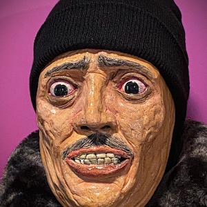 May include: A close-up of a realistic-looking mask with a surprised expression. The mask is made of brown and tan clay and has a black knit beanie on top. The mask has a mustache and a wide open mouth with visible teeth.