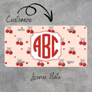 May include: A personalized license plate with a light pink background, adorned with red cherries and stars. The center features a red monogram within a white frame. The words "Customize" and "License Plate" are also visible.