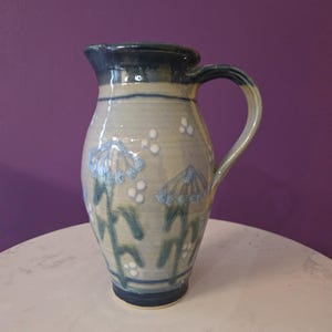 Könnte beinhalten: Keramikkrug mit blau-weißem Blumenmuster. Der Krug hat einen Henkel und einen Ausguss. Die Blumen sind blau mit weißen Akzenten. Der Krug steht auf einer weißen Oberfläche mit einem violetten Hintergrund.