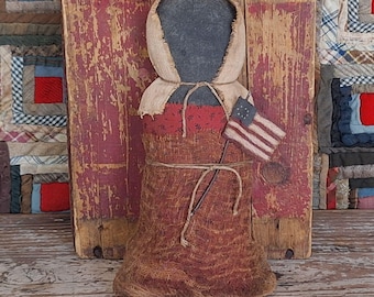 Muñeca primitiva negra de la pradera con vestido de calico de reproducción y delantal desgastado con bandera estadounidense pintada.