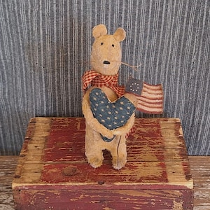 May include: A tan-colored mouse figurine holding a blue heart with white stars and a small American flag. The mouse wears a red and white plaid scarf. The figurine is placed on a rustic, red and brown wooden box.