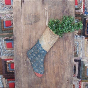 May include: A fabric stocking featuring a blue section with a white circular pattern and a tan upper cuff. The stocking has a red toe and is filled with green artificial pine needles, hanging from a simple string loop against a wooden board.