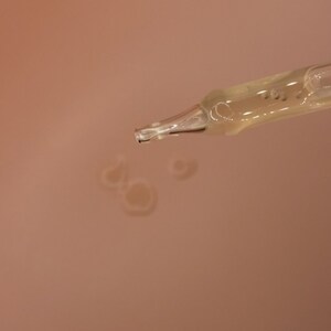 Op de afbeelding: Close-up van een heldere glazen pipet die een gele vloeistof afgeeft. De pipet is schuin, met druppels van de vloeistof die zich vormen op een perzik-kleurig oppervlak. De afbeelding benadrukt de toepassing van huidverzorgings- of cosmetische producten.