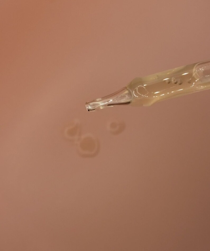 Op de afbeelding: Close-up van een heldere glazen pipet die een gele vloeistof afgeeft. De pipet is schuin, met druppels van de vloeistof die zich vormen op een perzik-kleurig oppervlak. De afbeelding benadrukt de toepassing van huidverzorgings- of cosmetische producten.