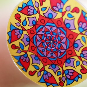 May include: A colorful mandala design painted on a round surface. The design features red, blue, yellow, purple, and white colors. The mandala is surrounded by a border of red and blue flowers.