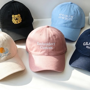 May include: Five baseball caps in black, light blue, cream, dusty pink, and navy blue. The black cap features a yellow tiger head, the light blue has white coordinates, the cream has a shell and sun, the pink says Embroidery Custom, and the navy says GRANDPA EST. 2025.