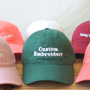 Può includere: Cinque cappellini da baseball di diversi colori con testo ricamato. I cappellini sono rosa, verde, bianco, rosso e arancione. Il testo sui cappellini recita "Custom Embroidery", "Saving Grace" e "Go Easy".