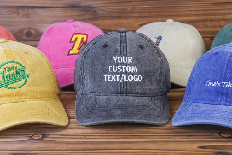 May include: Six different colored baseball caps with various embroidered designs. The caps are made of denim and have adjustable straps. The caps are arranged in a row on a wooden surface. One cap has the text "YOUR CUSTOM TEXT/LOGO" embroidered on it.