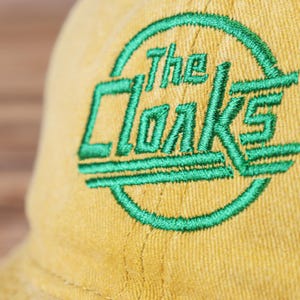 May include: A yellow baseball cap with a green embroidered logo that says "The Cloaks".