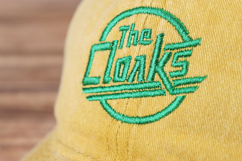 May include: A yellow baseball cap with a green embroidered logo that says "The Cloaks".