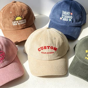 May include: Five corduroy baseball caps in brown, navy blue, pink, beige, and olive green. The brown cap says LET'S WATCH THE SUNSET, the navy cap says TODAY IS A GOOD DAY, the pink cap says I LOVE YOU, the beige cap says CUSTOM YOUR DESIGN, and the olive cap says BUTTERFLY FREE LIFE.