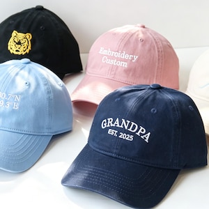 Puede incluir: Una colección de cinco gorras de béisbol en negro, azul claro, rosa, azul marino y crema. La gorra negra tiene una cabeza de tigre amarilla, la azul claro coordenadas blancas, la rosa dice Embroidery Custom, la azul marino GRANDPA EST. 2025 y la crema un sol amarillo y una concha azul.