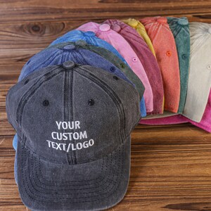 May include: A stack of 10 blank, customizable, cotton twill baseball caps in various colors, including blue, pink, yellow, orange, and gray. The top cap is dark gray with the text "YOUR CUSTOM TEXT/LOGO" embroidered in white.