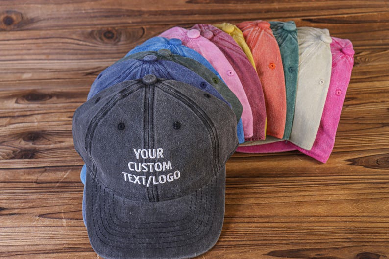 May include: A stack of 10 blank, customizable, cotton twill baseball caps in various colors, including blue, pink, yellow, orange, and gray. The top cap is dark gray with the text "YOUR CUSTOM TEXT/LOGO" embroidered in white.
