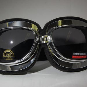 May include: Silver aviator goggles with black padding and dark gray lenses. The lenses are labeled "Shatterproof Polycarbonate" and "Polycarbonate Lens UV 400 100% Protection".