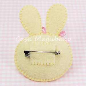 May include: A felt bunny brooch with pink ribbon accents and a safety pin closure. The bunny is cream colored with yellow stitching around the edges.