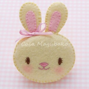 May include: A felt bunny with pink ears and a pink bow. The bunny has a stitched face with brown eyes and a pink nose.