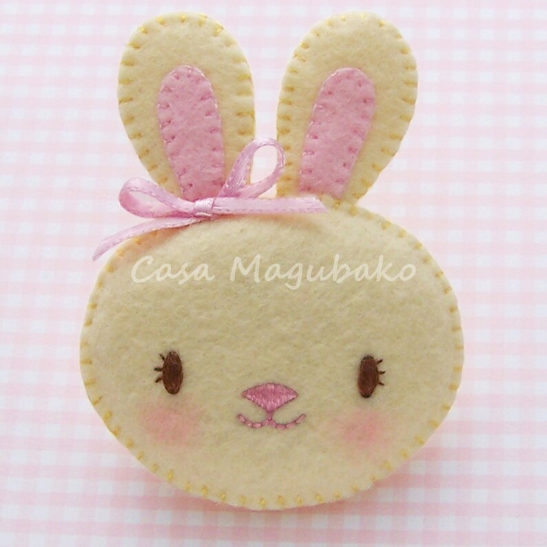 May include: A felt bunny with pink ears and a pink bow. The bunny has a stitched face with brown eyes and a pink nose.