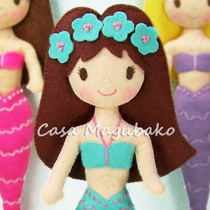 May include: A brown-haired felt doll with a blue and pink mermaid tail, wearing a turquoise top and a flower crown with blue and pink flowers. The doll has a smiling face with brown eyes.
