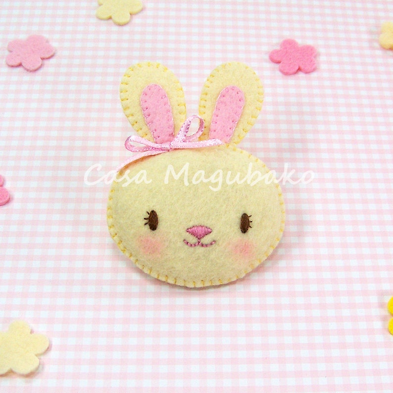 May include: A yellow felt bunny with pink ears and a pink bow. The bunny has embroidered eyes, a nose, and a mouth.
