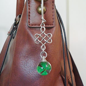 May include: A silver charm with a Celtic knot design and a clear glass sphere containing a four-leaf clover. The charm is attached to a brown leather handbag.