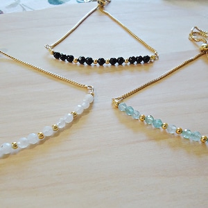 May include: Three gold chain bracelets with adjustable sliding knots. The bracelets feature different colored gemstone beads: black, white, and light green.