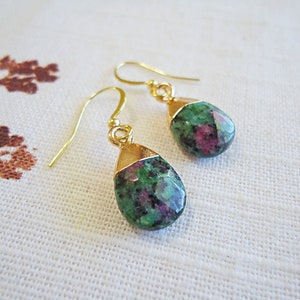 May include: A pair of gold-colored drop earrings. Each earring features a teardrop-shaped gemstone with a gold-colored cap. The gemstones are a mix of green and red hues. The earrings are displayed on a white surface.