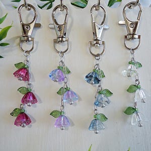 May include: Four keychains with silver clasps and dangling charms. Each charm features a cluster of small, clear glass bells with green leaves. The bells are in shades of pink, purple, blue, and clear.