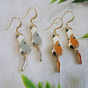 May include: A pair of gold cat earrings with a dangling tail. One pair features a grey, white, and black cat, while the other pair features an orange, white, and black cat.