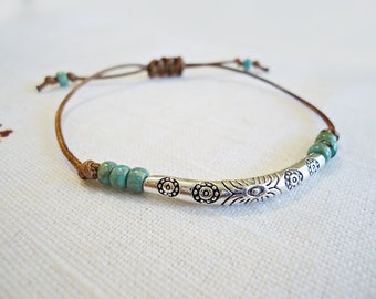 May include: A bracelet featuring a brown cord with an adjustable sliding knot closure. The center displays a curved silver-toned metal tube with etched circular patterns, flanked by three small turquoise-colored beads on each side.