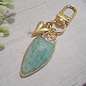 May include: Gold-toned keychain with a heart charm and a teardrop-shaped pendant. The pendant features a light blue-green stone with a gold border. The keychain has a clasp for easy attachment.