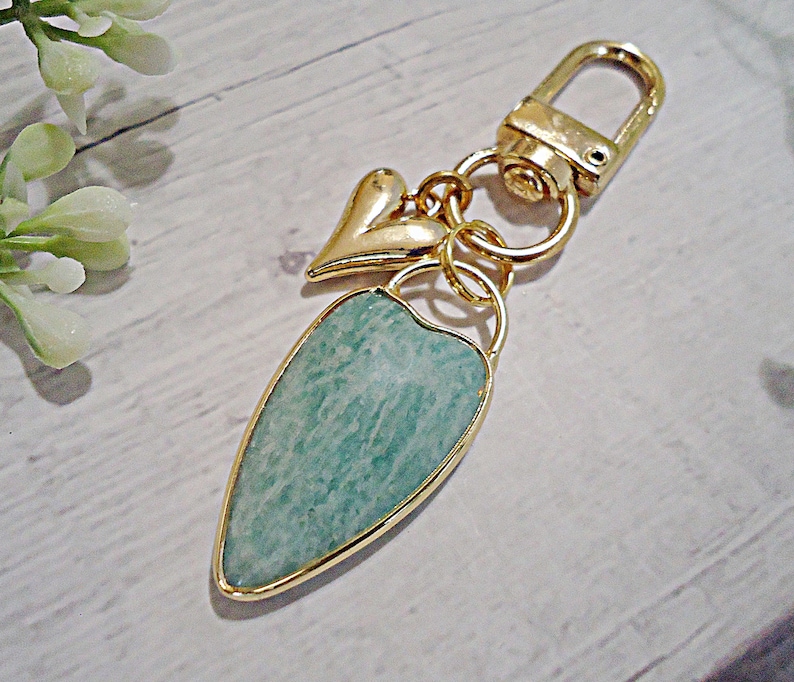 May include: Gold-toned keychain with a heart charm and a teardrop-shaped pendant. The pendant features a light blue-green stone with a gold border. The keychain has a clasp for easy attachment.