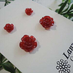 May include: Three red rose-shaped resin embellishments on a white background. The background has the text "Lanza Creations" in black.
