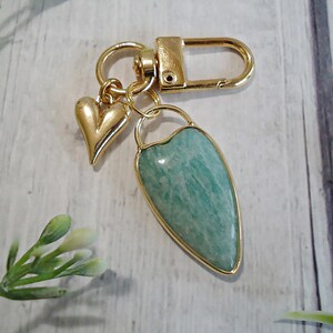 May include: A gold-colored keychain with a heart charm and a teardrop-shaped pendant. The pendant features a light green stone with a gold-colored border. The keychain has a clasp for easy attachment.