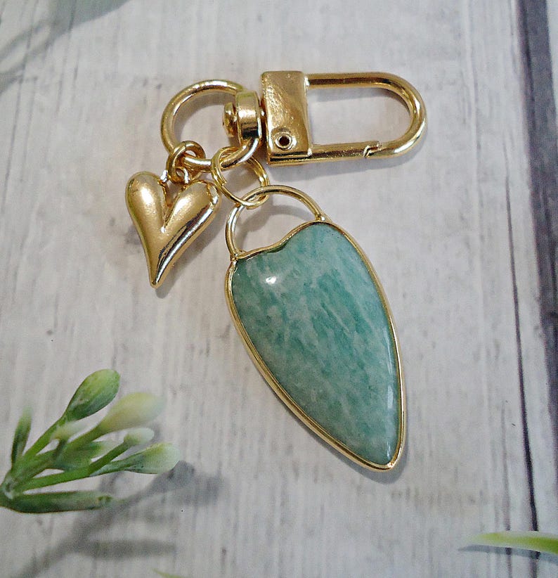 May include: A gold-colored keychain with a heart charm and a teardrop-shaped pendant. The pendant features a light green stone with a gold-colored border. The keychain has a clasp for easy attachment.