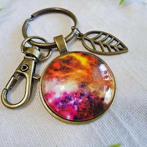 May include: A bronze key chain with a round glass charm featuring a colorful nebula image. The charm is attached to a split ring with a bronze leaf charm.