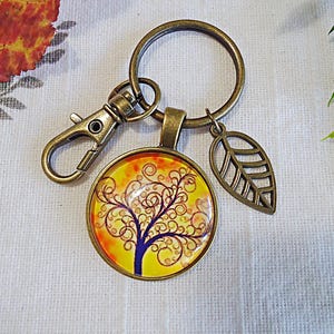 May include: A bronze key chain with a round glass charm featuring a purple tree with yellow and orange swirls on a yellow background. The charm is attached to a bronze ring with a bronze leaf charm.