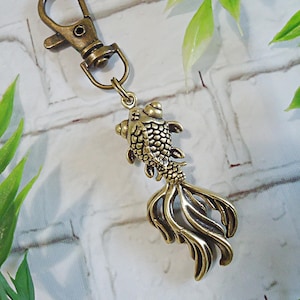 May include: A bronze-colored keychain featuring a detailed goldfish charm. The fish has textured scales and flowing fins, attached to a clasp. The background is a white brick wall with green foliage.