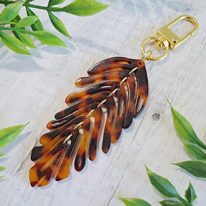 May include: A keychain featuring a brown and black tortoiseshell patterned acrylic leaf charm. The leaf is composed of multiple articulated segments connected by small gold-toned metal rings, attached to a gold-toned lobster claw clasp.