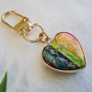 May include: Gold heart-shaped keychain with a multi-colored stone inlay. The stone has stripes of green, blue, yellow, orange, and red.